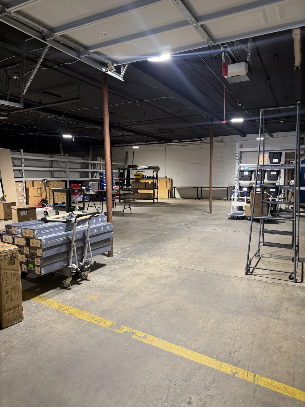 An open warehouse floor with shelving, boxes, a rolling ladder and a cart of flat-packed furniture