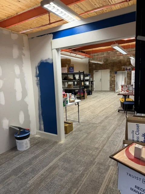 An office mid-renovation with patched drywall, a partly painted blue wall, a paint bucket and shelving in the room beyond